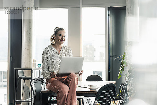 Lächelnde Geschäftsfrau sitzt im Büro mit Laptop auf dem Tisch