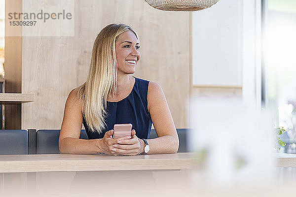 Porträt einer lächelnden blonden Geschäftsfrau mit Smartphone  die in einem Café wartet