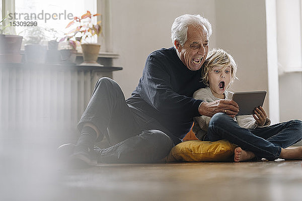 Schreiender Großvater und Enkel  die zu Hause auf dem Boden sitzen und ein Tablett benutzen