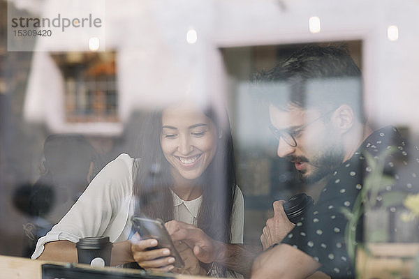 Junges Paar mit Smartphone hinter einer Fensterscheibe in einem Café