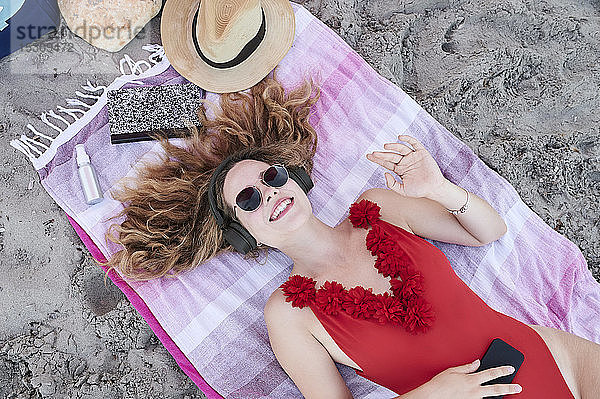 Glückliche junge Frau liegt auf einem Handtuch am Strand und hört Musik