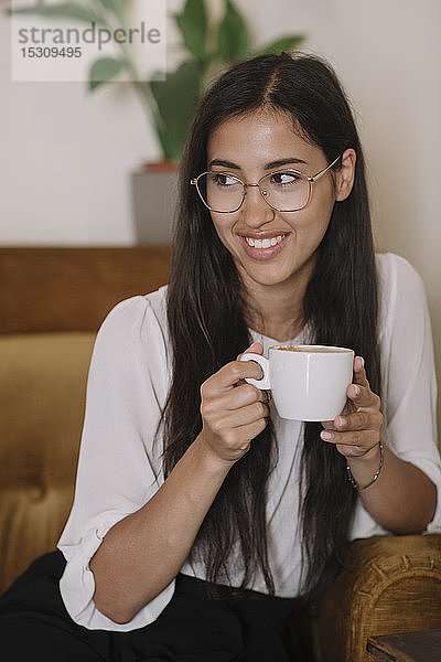 Lächelnde junge Frau mit Tasse Kaffee im Sessel eines Cafés sitzend