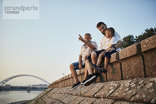 Großvater  Vater und Sohn sitzen auf einer Mauer am Flussufer