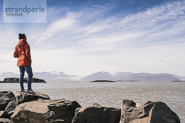 Junge Frau steht auf Felsblöcken und schaut auf das Meer  Südost-Island