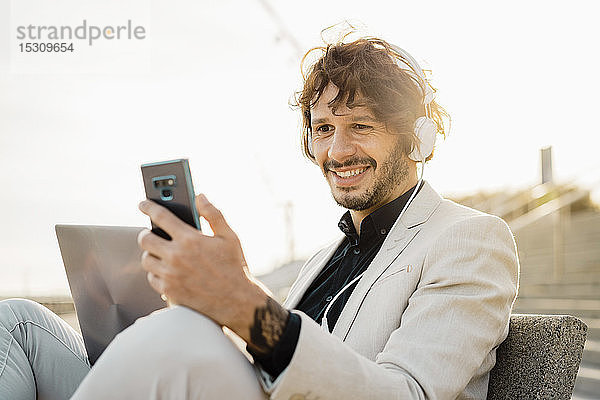 Porträt eines lächelnden Geschäftsmannes mit Kopfhörer und Laptop  der auf sein Mobiltelefon schaut