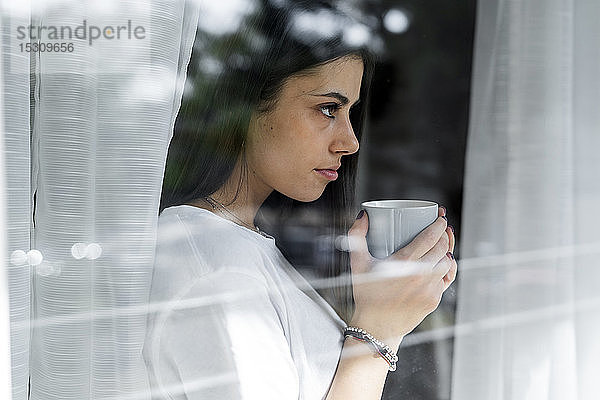 Junge Frau mit Tasse Kaffee hinter Fensterscheibe