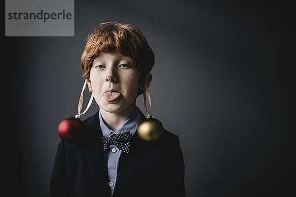 Porträt eines rothaarigen Jungen mit Fliege und Weihnachtskugeln mit herausgestreckter Zunge