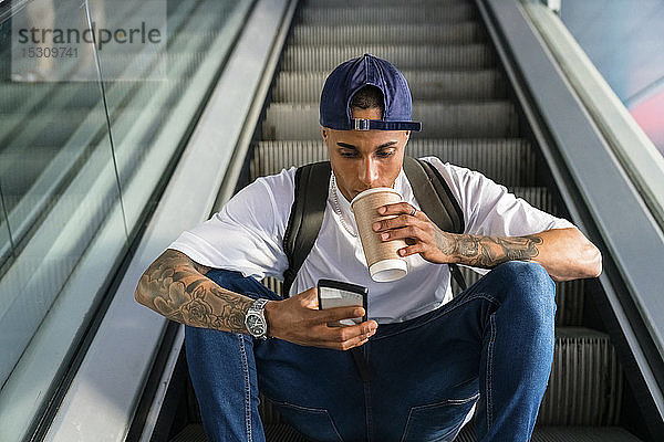 Junger Mann mit Rucksack sitzt auf der Rolltreppe und schaut auf sein Handy  während er Kaffee zum Mitnehmen trinkt