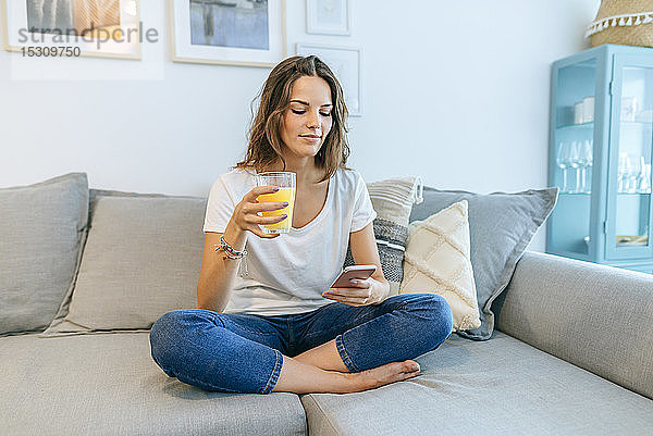 Junge Frau sitzt mit Saft auf dem Sofa und telefoniert mit ihrem Handy