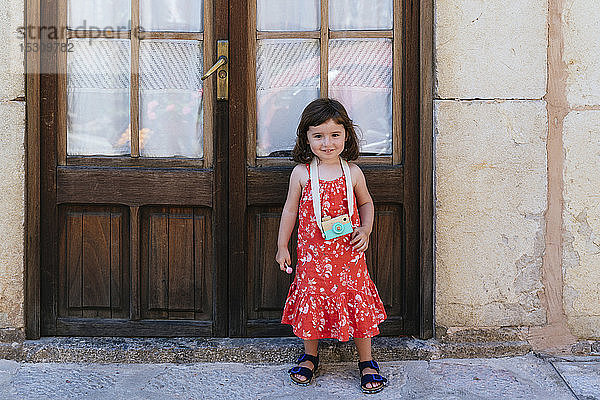Porträt eines lächelnden kleinen Mädchens mit Holzspielzeugkamera in rotem Kleid mit Blumenmuster