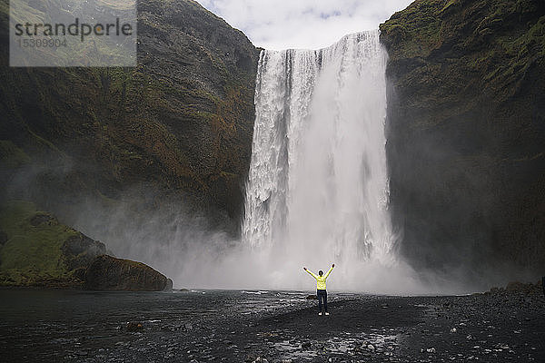Junge Frau steht mit erhobenen Armen vor den Skogafoss-Wasserfällen  Island