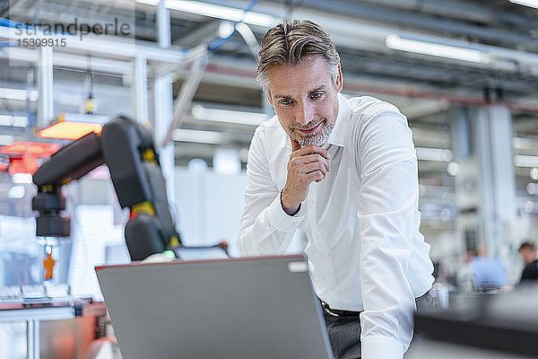 Selbstbewusster Geschäftsmann in einer modernen Fabrikhalle mit Laptop