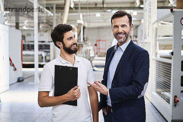 Porträt eines lächelnden Geschäftsmannes und Angestellten mit Klemmbrett in einer Fabrik