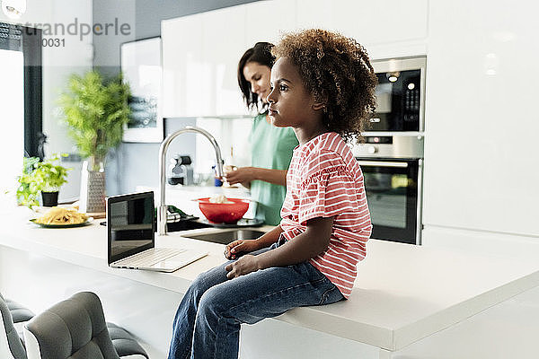 Mädchen sitzt auf dem Küchentisch mit Laptop und Mutter im Hintergrund