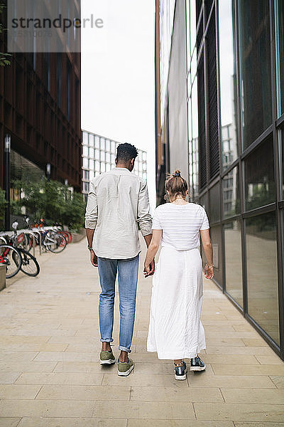 Rückenansicht eines jungen Paares  das Hand in Hand auf dem Bürgersteig geht