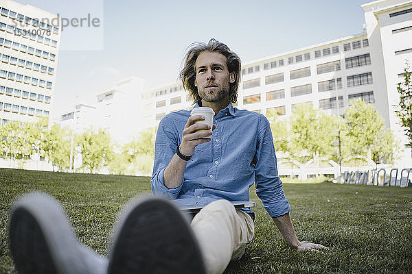Junger Mann sitzt auf einer Wiese mit Kaffee zum Mitnehmen