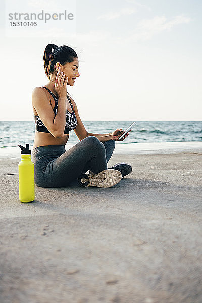 Frau benutzt Smartphone und In-Ohr-Gerät während des Trainings und sitzt auf einem Pier