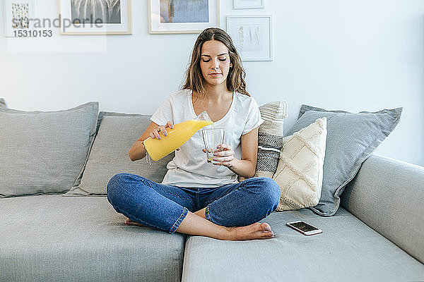 Junge Frau sitzt auf einem Sofa und gießt Orangensaft in ein Glas