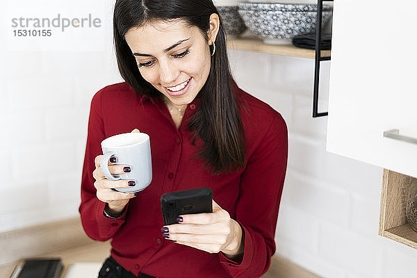 Lächelnde junge Geschäftsfrau mit Handy und Tasse Kaffee zu Hause