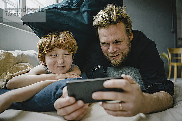Porträt von Vater und Sohn  die auf der Couch liegen und auf ein Handy schauen