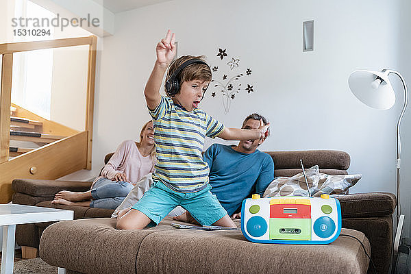 Aufgeregter Junge  der mit Kopfhörern auf der Couch zu Hause mit den Eltern im Hintergrund Musik hört