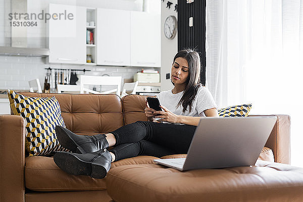 Junge Frau sitzt zu Hause auf der Couch und benutzt Laptop und Handy