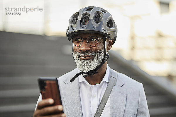 Porträt eines lächelnden reifen Geschäftsmannes mit Smartphone  der einen Fahrradhelm und eine Brille trägt