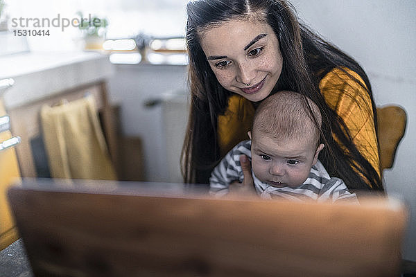 Mutter mit Baby mit Laptop auf dem Küchentisch