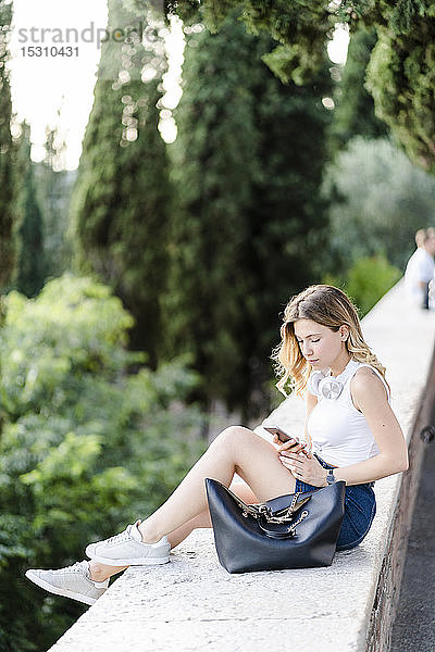 Junge Frau mit Tasche  die mit einem Smartphone an einer Wand sitzt