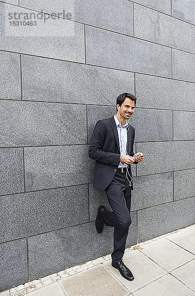 Lächelnder Geschäftsmann lehnt mit Kopfhörern und Mobiltelefon an Wand
