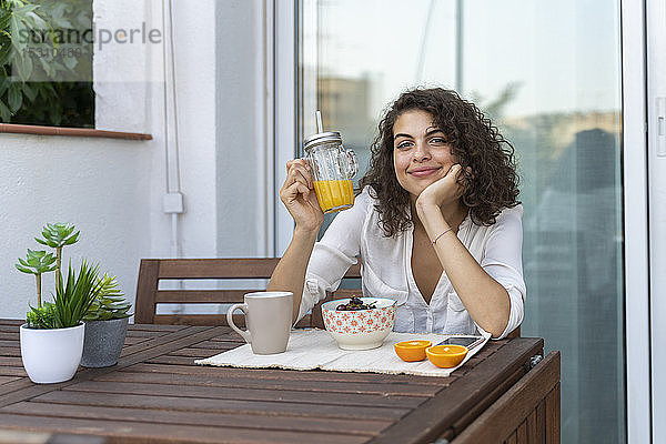 Porträt einer lächelnden jungen Frau mit Orangensaft auf dem Balkon
