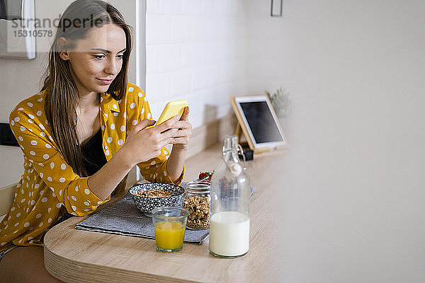 Junge Frau benutzt Mobiltelefon und frühstückt zu Hause in der Küche