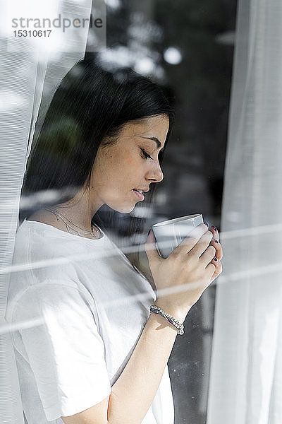 Junge Frau mit Tasse Kaffee hinter Fensterscheibe