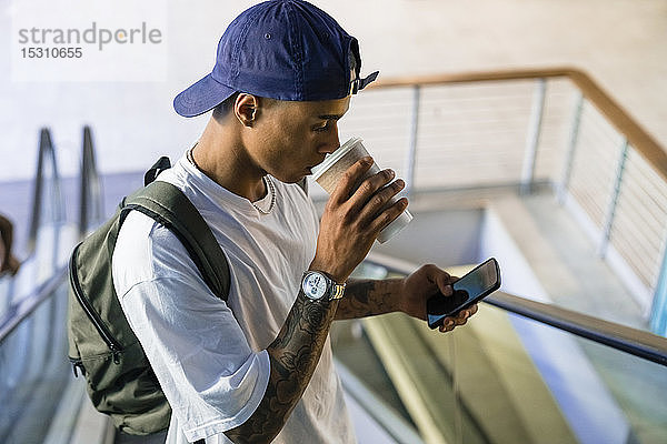 Tätowierter junger Mann mit Rucksack steht auf Rolltreppe und schaut auf Handy  während er Kaffee zum Mitnehmen trinkt