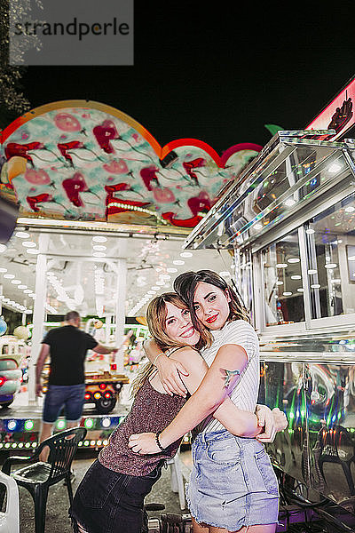 Porträt von zwei glücklichen jungen Frauen  die sich nachts auf einem Jahrmarkt umarmen