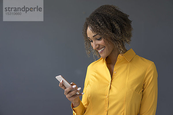 Porträt einer lächelnden jungen Frau mit gelbem Hemd  die auf ihr Handy schaut