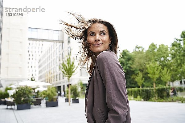 Lächelnde Geschäftsfrau mit windgepeitschtem Haar in der sich umdrehenden Stadt