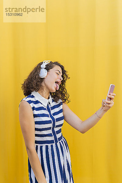 Porträt einer jungen Frau mit Kopfhörern  die ein Smartphone benutzt  ihre Zunge herausstreckt und ein Selfie macht