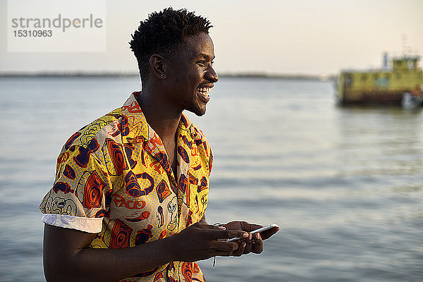 Junger Mann steht am Meer  hält Smartphone  Porträt