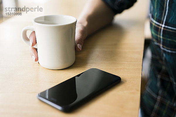 Hand einer jungen Frau  die eine Tasse Kaffee neben einem Smartphone hält