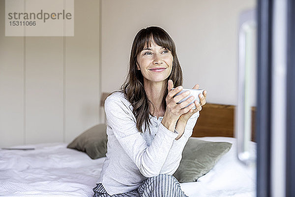 Entspannte Frau mit Tasse Kaffee sitzt zu Hause auf dem Bett
