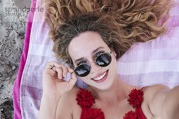 Selbstporträt einer lächelnden jungen Frau  die auf einem Handtuch am Strand liegt