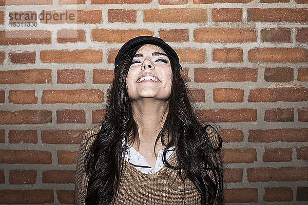 Porträt einer lächelnden jungen Frau mit geschlossenen Augen vor einer Ziegelmauer