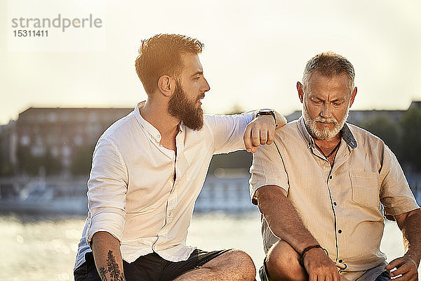 Vater und erwachsener Sohn sitzen auf einer Mauer am Flussufer und unterhalten sich