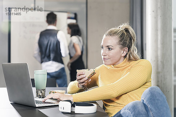 Lässige Geschäftsfrau  die im Büro mit Laptop am Tisch sitzt und ein Sandwich isst