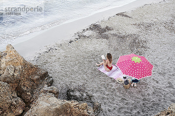 Junge Frau sitzt auf einem Handtuch am Strand und liest ein Buch