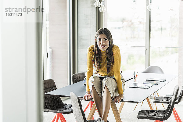 Porträt einer selbstbewussten jungen Geschäftsfrau  die im Büro auf dem Tisch sitzt