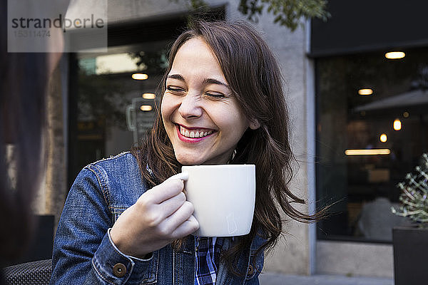 Fröhliche junge Frau lacht mit geschlossenen Augen und hält Kaffeetasse im Außencafé in Madrid  Spanien