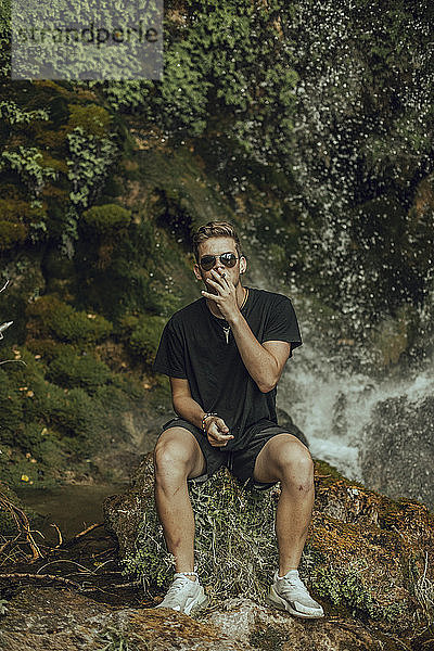 Porträt eines coolen jungen Mannes an einem Wasserfall  der einen Joint Marihuana raucht