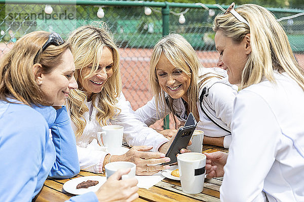 Gruppe von Frauen  die Tee und Kaffee mit Keksen geniessen und sich nach einem Spiel im Tennisclub ein Smartphone teilen